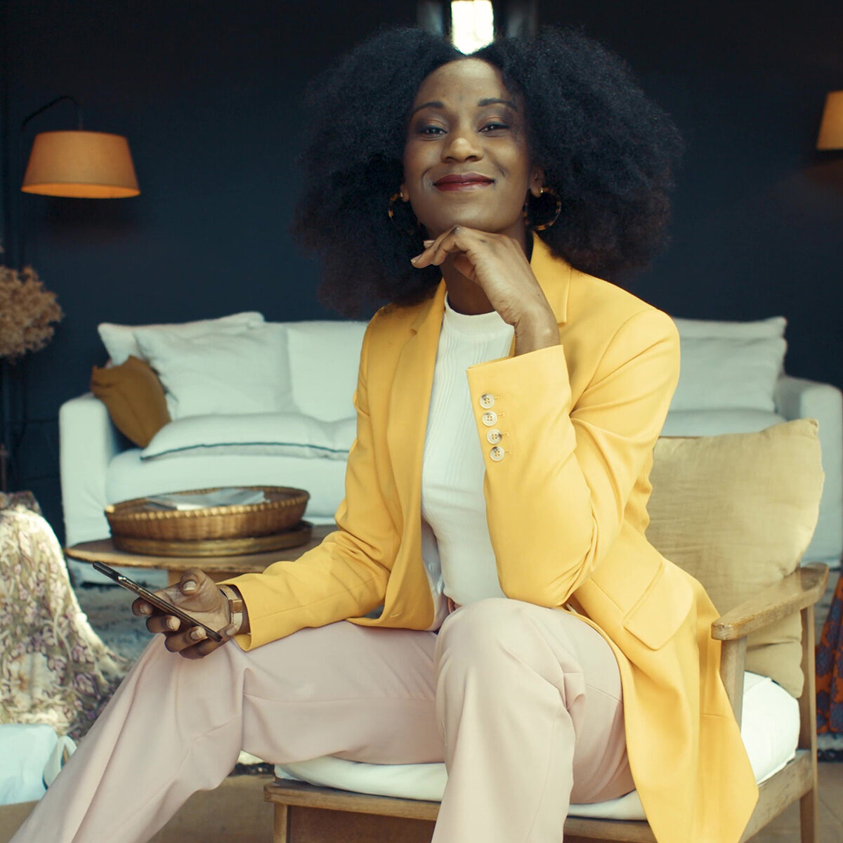 Spot publicitaire MYM : femme souriante en blazer jaune tenant un smartphone dans un salon cosy