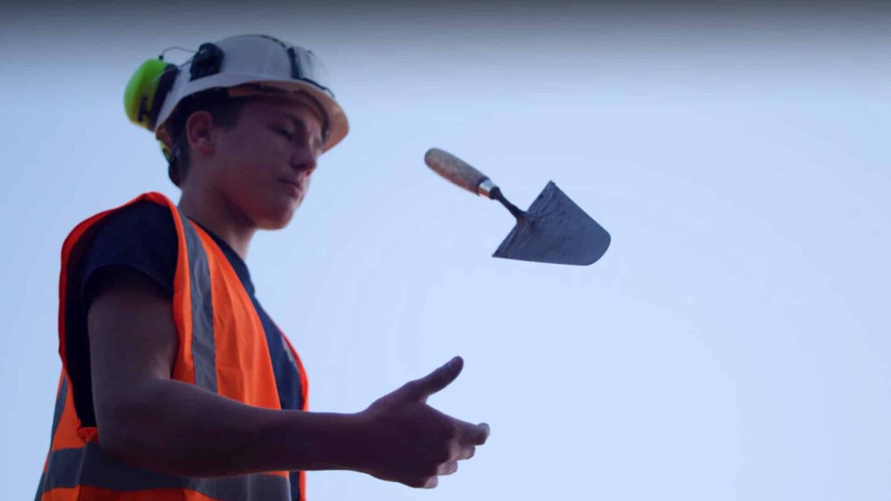 Jeune ouvrier BTP en gilet orange faisant tournoyer une truelle, film d'onboarding Médinger travaux publics